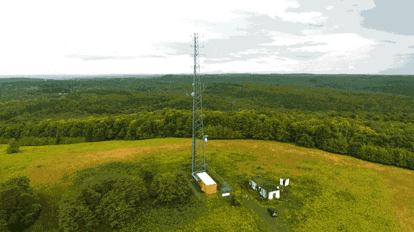 A short clip of video of a radio tower from the air. The tower sits at the top of a hill surrounded by green fields and forrest. There are several small buildings and a car at the base of the tower.