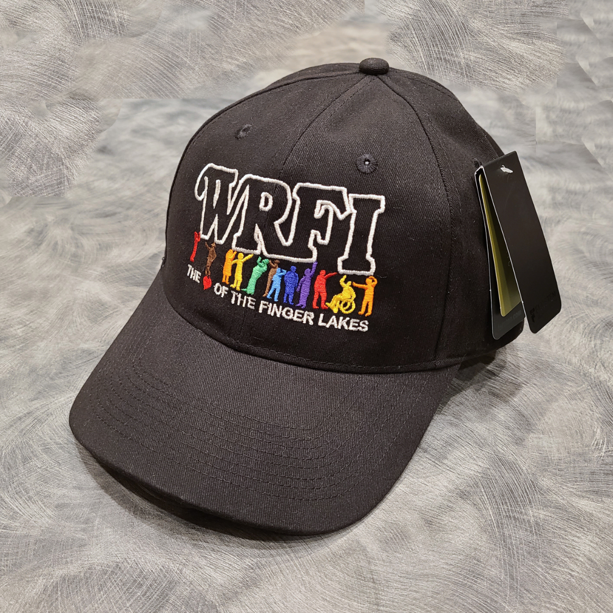 A black baseball cap on a brushed grey tabletop. The image on the front of the hat features the letters WRFI being help up by a groups of small figures in a range of colors. One figure sits in a wheelchair.