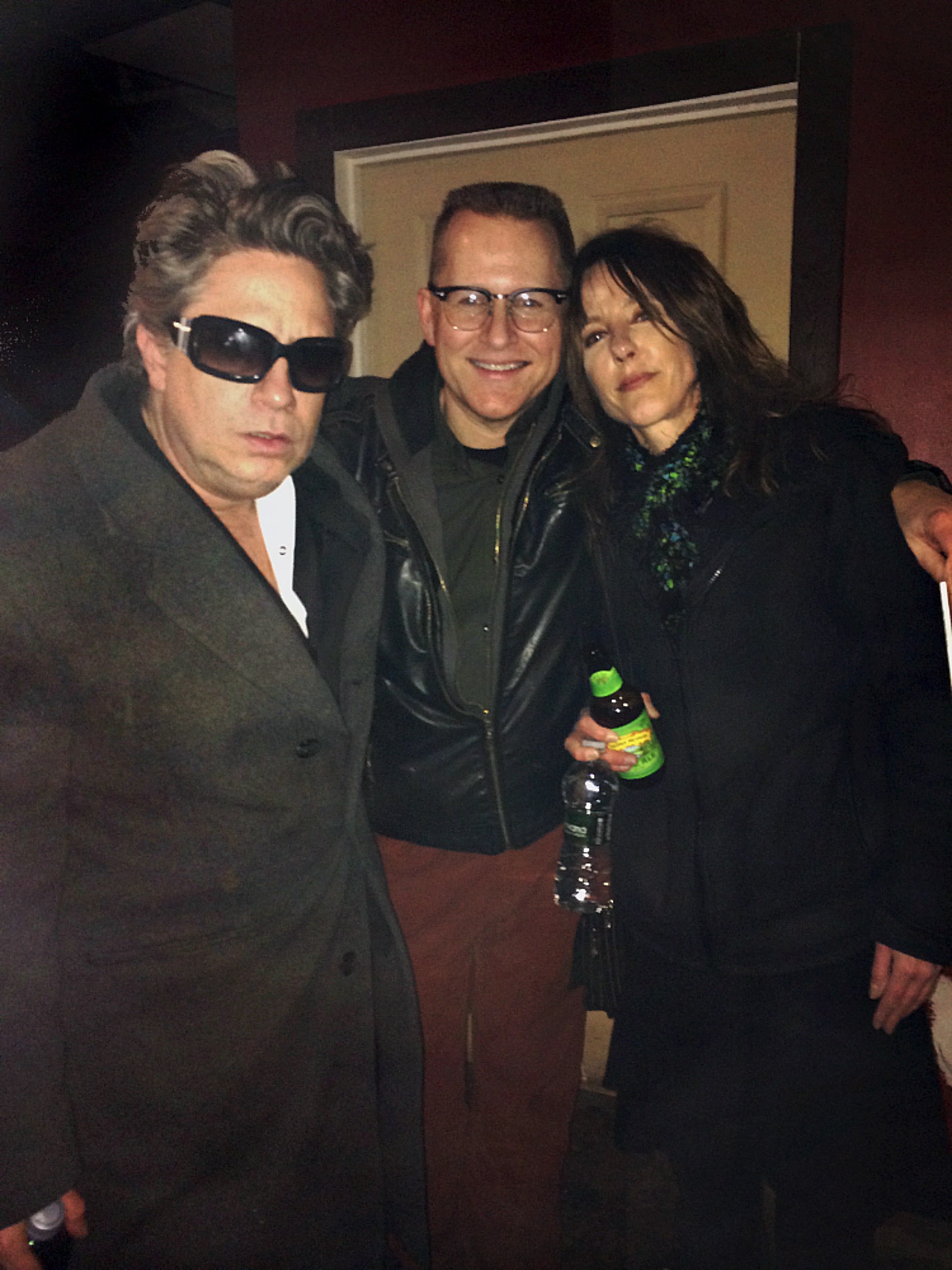 Dexter Romweber (left) a white man with greying, medium length, dark hair in dark overcoat and sunglasses, Rocket Morton (center) a white man smiling in a leather jacket with a crew cut, and Sara Romweber (right) a white woman with long hair in an overcoat and green sparkling scarf holding a beer and a water bottle.