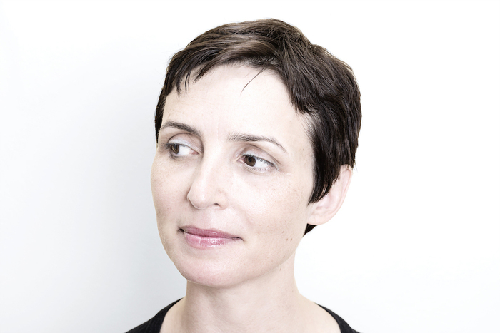 A white woman with dark short cropped hair looks to the left of the screen. The background is stark white, and you can just see the collar of a black shirt.