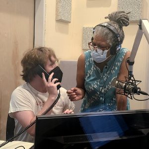 Two people are talking in a radio recording booth. One is a young white man wearing a white shirt, and a black face mask holding a black corded telephone. The other is an older black woman with grey locs and a blue patterned tanktop and totoiseshell glasses.