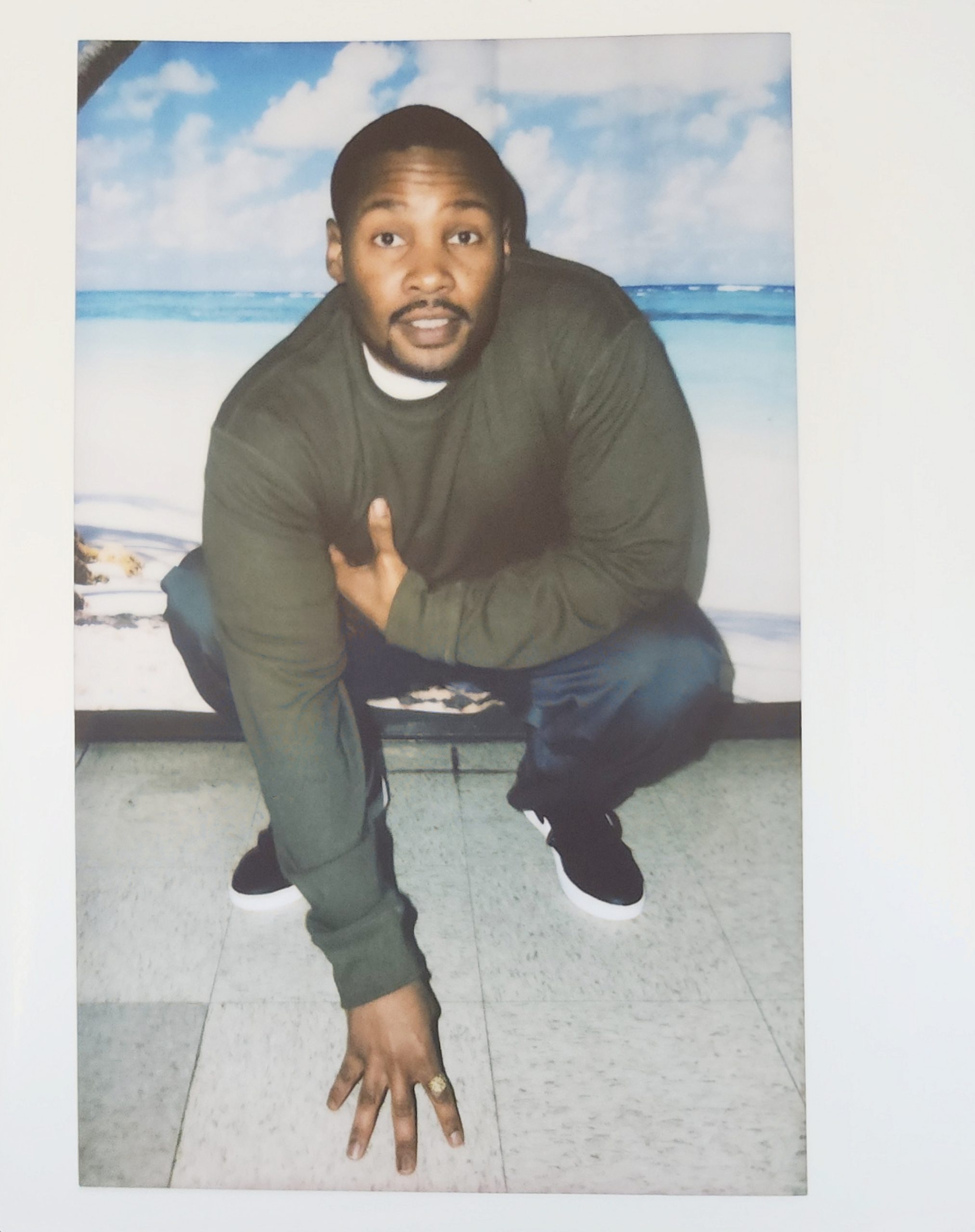 Image of a young black man with a short cropped goatee, and short hair wearing a dark green long sleeve shirt and navy pants, crouched in front of a backdrop showing a beach on a sunny day. the man looks directly at the camera with a relaxed expression