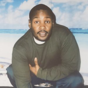 Image of a young black man with a short cropped goatee, and short hair wearing a dark green long sleeve shirt and navy pants, crouched in front of a backdrop showing a beach on a sunny day. the man looks directly at the camera with a relaxed expression