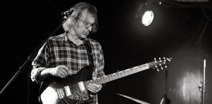 black and white photograph of a man playing guitar. The man stands just behind a microphone wearing glasses, he holds an electric guitar.