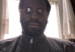 A black man with round glasses is seated in front of a bright window with white curtains.