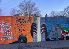 A large hand-painted sign in dusk light. Images include black hands on bars, minarets topped with crescent moons, planes dropping bombs, a man holding a bloodied child, and a slice of watermellon. The sign reads, "Over 35,000 killed, over 14,000 of whom are children," "The sun will shine on a free Palestine," and "Land Back."