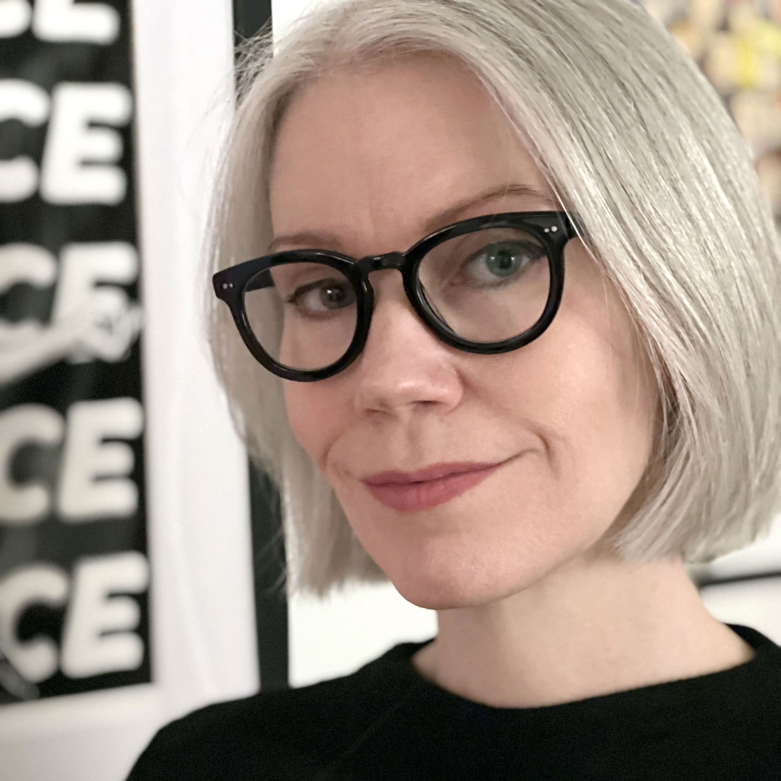 A white woman with white hair cut in a short bob wears black rimmed glasses looks into the camera with her head tilted slightly