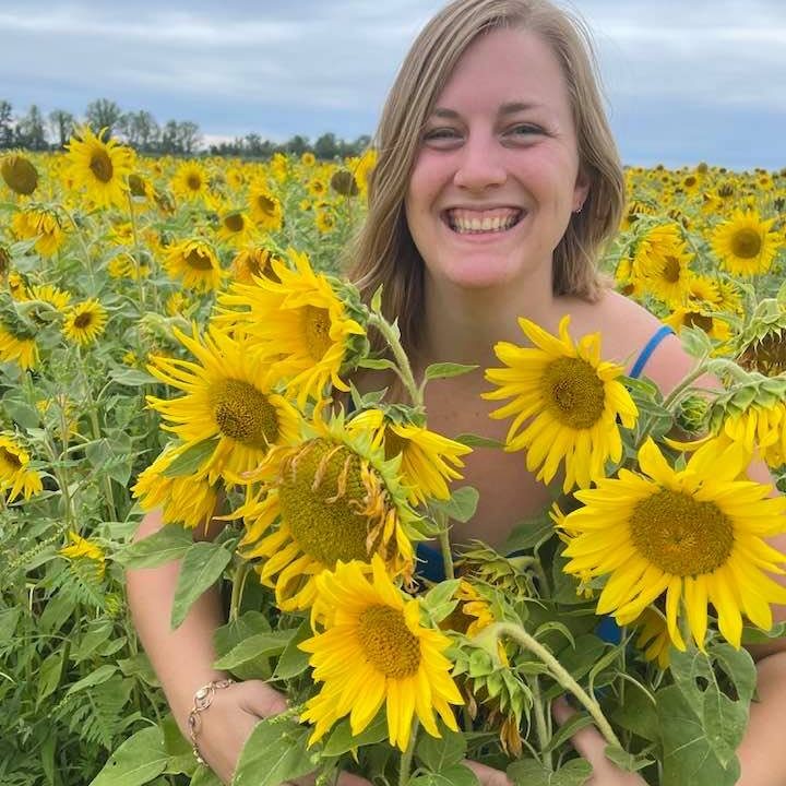 a blonde woman stands in a field of yellow sunflowers smiling into the camera.